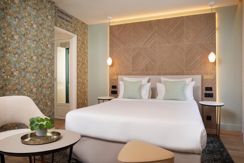 gay hotels in Paris: Modern hotel room with a large bed adorned with crisp white linens and mint green pillows, an intricate wooden headboard, and vintage floral wallpaper. The room is finished with a sleek side table, a stylish curved chair, and a potted plant, offering a blend of contemporary design and classic elegance.
