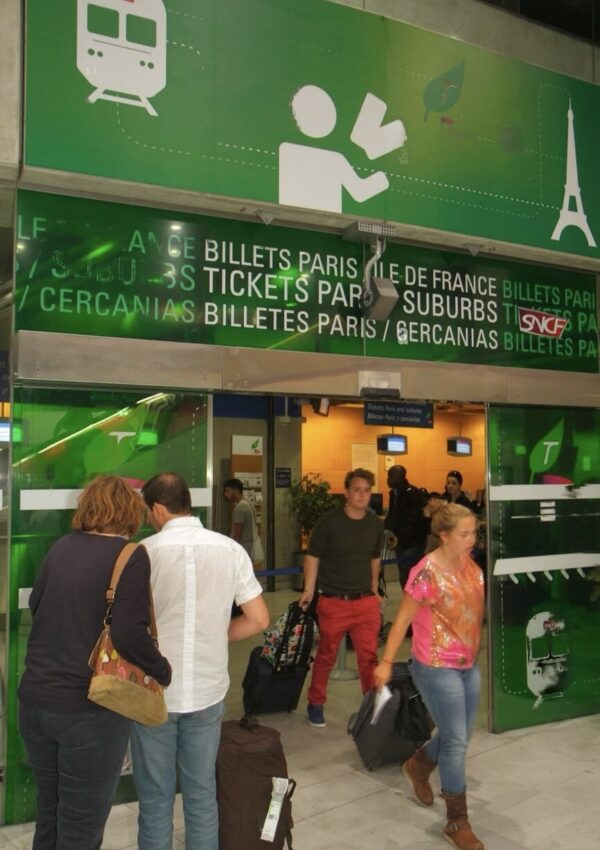 Travelers queuing at a ticket counter with 'Billets Paris Ile de France' signage overhead, indicating a train ticket purchase point for local and regional travel in Paris. The green-themed decor features pictograms of a train and a person receiving a ticket, along with the iconic Eiffel Tower, conveying a busy transport hub atmosphere.