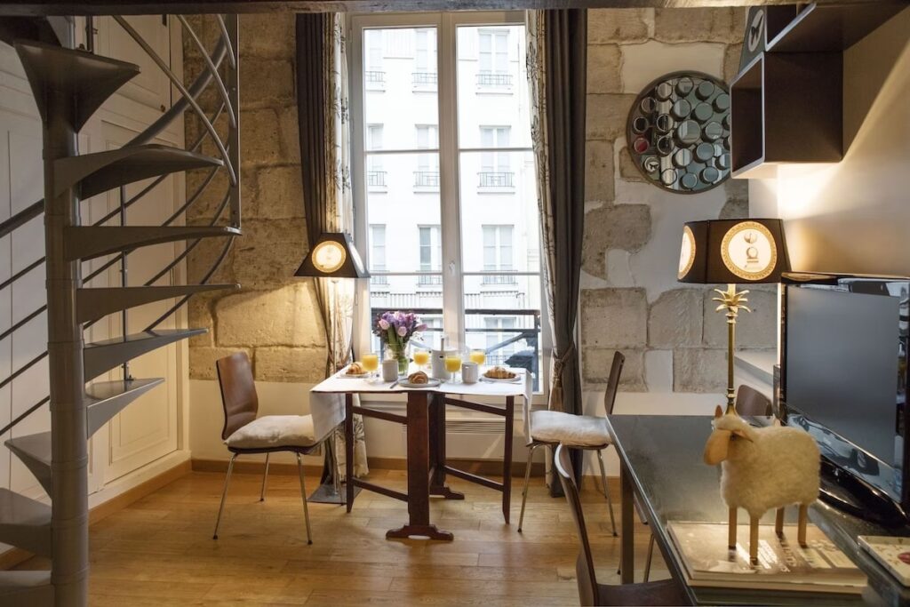 Chic dining area within a duplex studio, showcasing a rustic stone wall, a whimsical sheep sculpture, an intimate table set for breakfast with fresh juice and pastries, all under the warm glow of stylish lighting fixtures, with a view of Parisian buildings through the window.