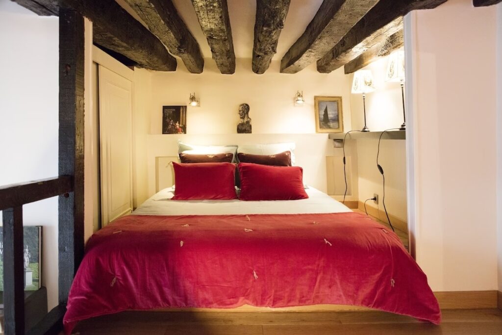 Romantic bedroom in a duplex studio with exposed wooden beams, a sumptuous bed with rich red linens, complemented by soft lighting and artistic decor, offering a cozy retreat in a classic Parisian setting.