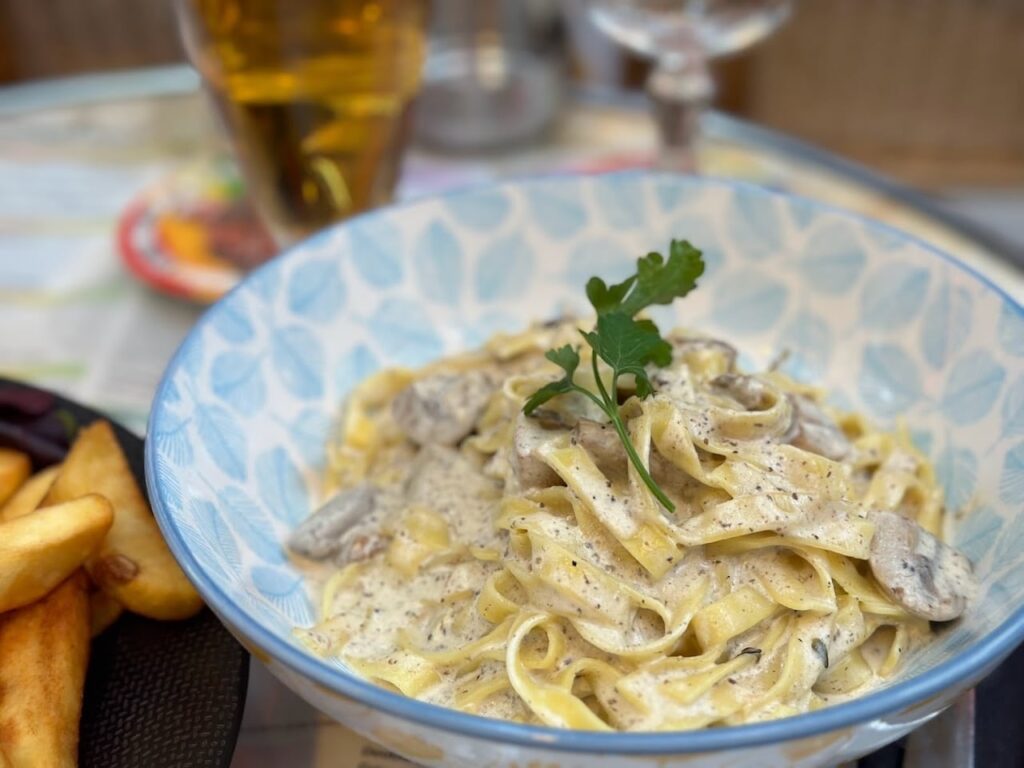Close-up of a bowl of creamy fettuccine pasta with mushrooms garnished with parsley, served at Le Ju' Restaurant in Paris, accompanied by golden fries and a refreshing glass of beer in the background.