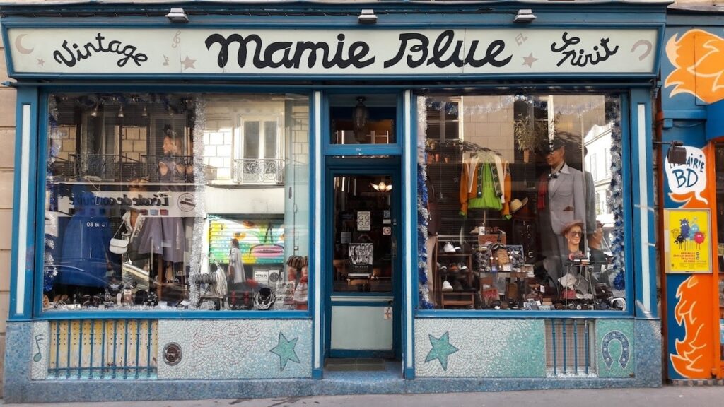 Vintage Shops in Paris: The quaint storefront of Mamie Blue, a vintage shop with a striking blue facade, displays a variety of retro clothing and accessories in its windows under a playful handwritten sign, inviting passersby into the world of vintage fashion.