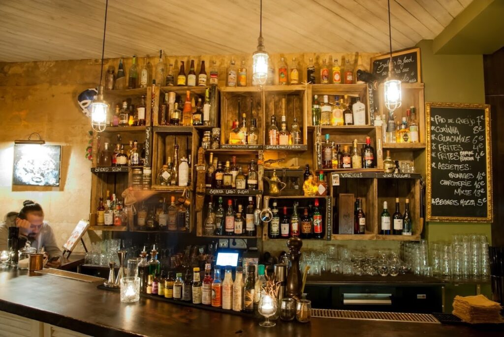 The rustic charm of Maria Loca bar in Paris, with its wooden shelves stocked with an eclectic selection of spirits and bottles, edison bulb lighting, and a chalkboard menu featuring cocktails and snacks.