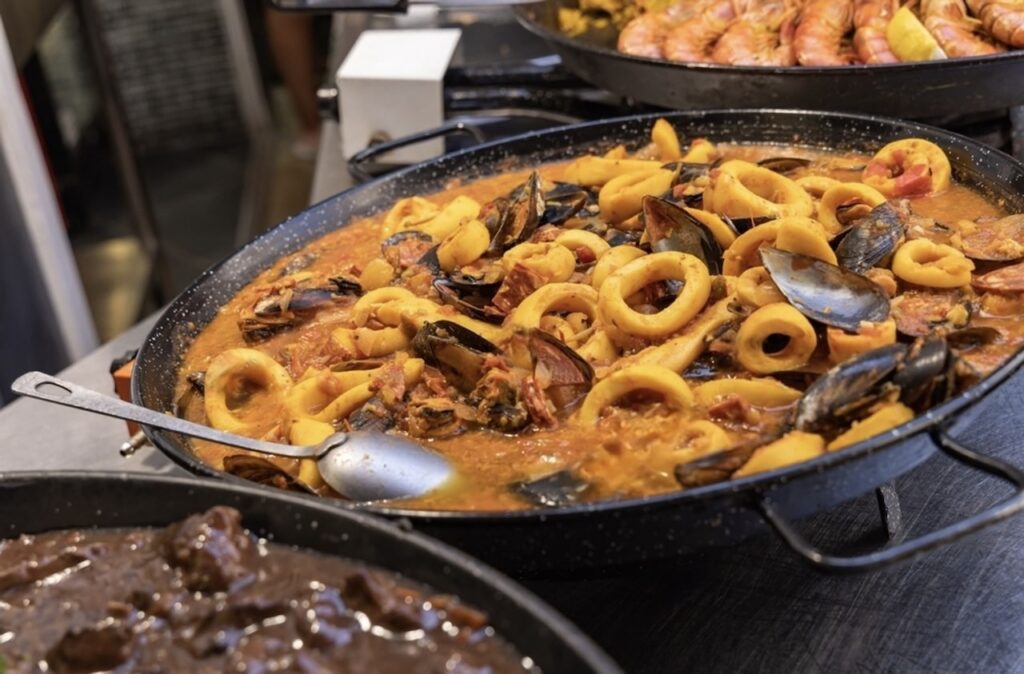 Marseille Food Guide: A large paella pan filled with seafood stew, featuring squid rings, mussels, and tomato-based sauce, showcased in a market setting with other dishes in the background, ready to be served to hungry patrons.