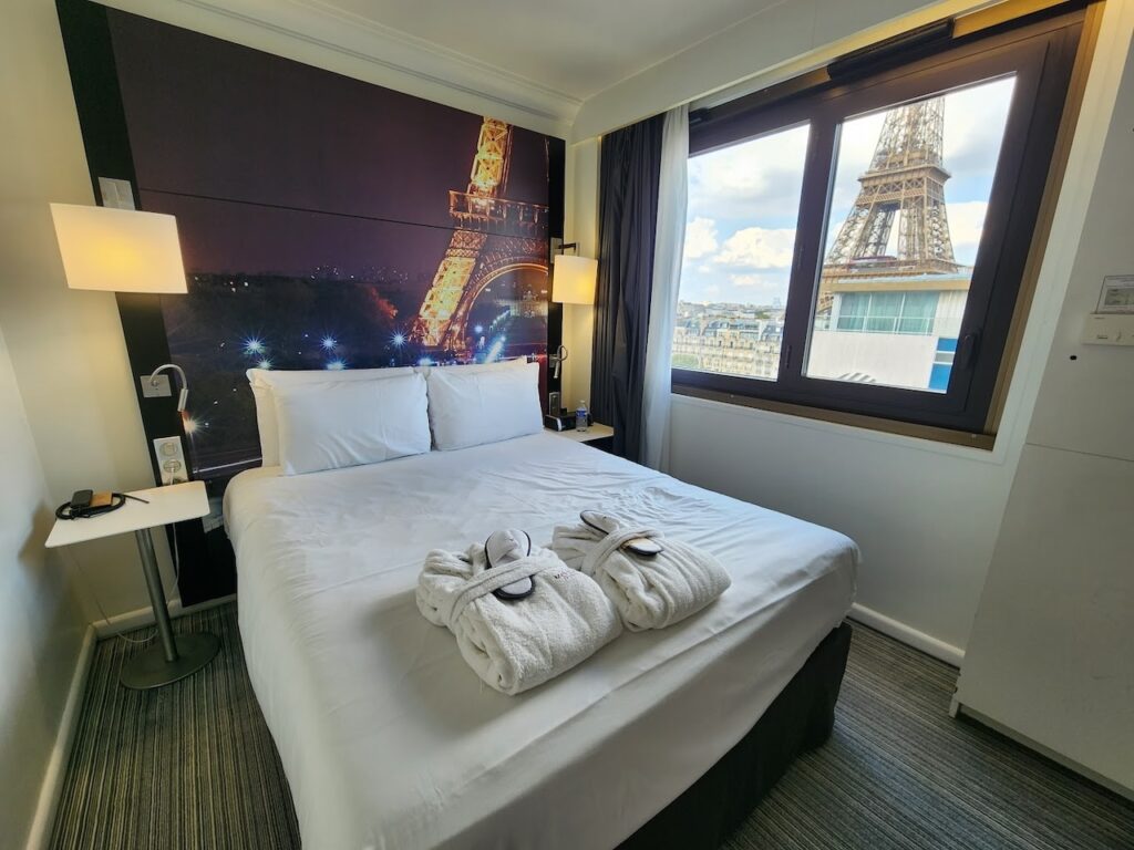 gay hotels in Paris: Hotel room with a panoramic view of the Eiffel Tower, both from the window and captured in a large night-time scene headboard photograph. The room is outfitted with a comfortable bed with white linens and towels folded into swan shapes, emphasizing the romantic and iconic Parisian experience.
