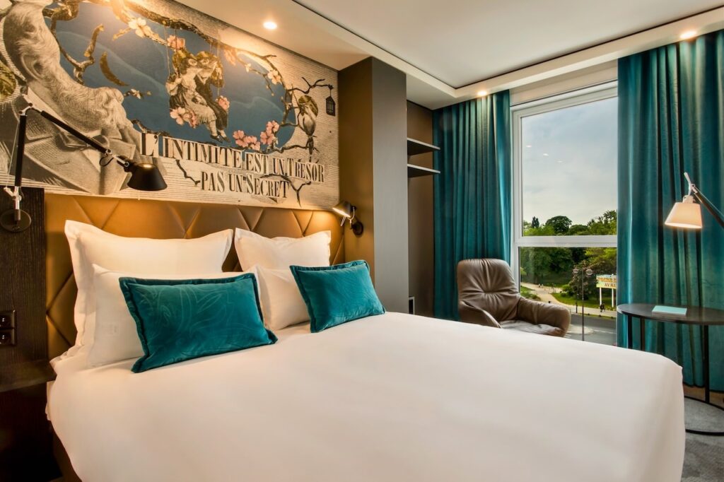 gay hotels in Paris: A luxurious hotel room with a large bed, white and teal pillows against a tufted headboard, and a dramatic wall art piece with classical imagery and the French phrase 'L'intimité est un trésor - pas un secret'. The room combines modern comfort with artistic flair, featuring a plush leather chair and teal curtains that frame a serene park view.