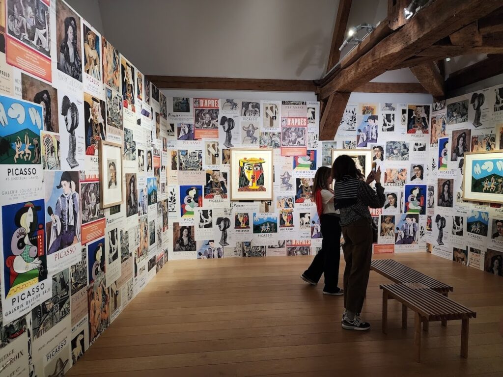 Visitors admiring a wall covered in a diverse collection of Picasso exhibition posters at the Musée Picasso in Paris, showcasing the extensive influence and variety of the artist's works in a room with exposed wooden beams.