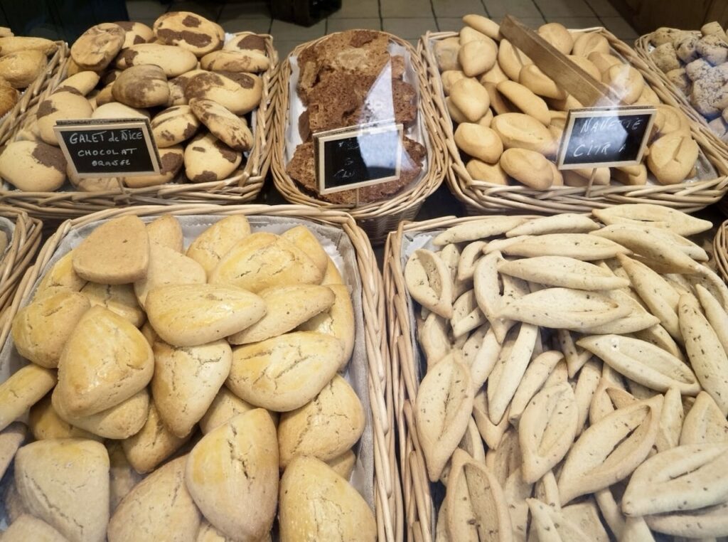 Marseille Food Guide: A variety of freshly baked breads displayed in wicker baskets, including chocolate and raisin galettes, rustic loaves, and navette cookies, each labeled with a chalkboard sign, offering a glimpse into a traditional bakery's assortment.
