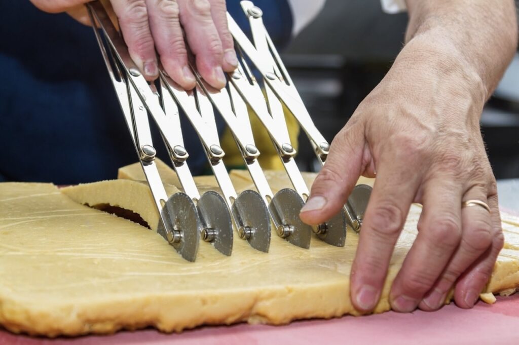 Marseille Food Guide: Close-up of a baker's hands using a multi-wheel pastry cutter to slice even strips of dough on a pink cutting board, demonstrating the precision required in pastry making.