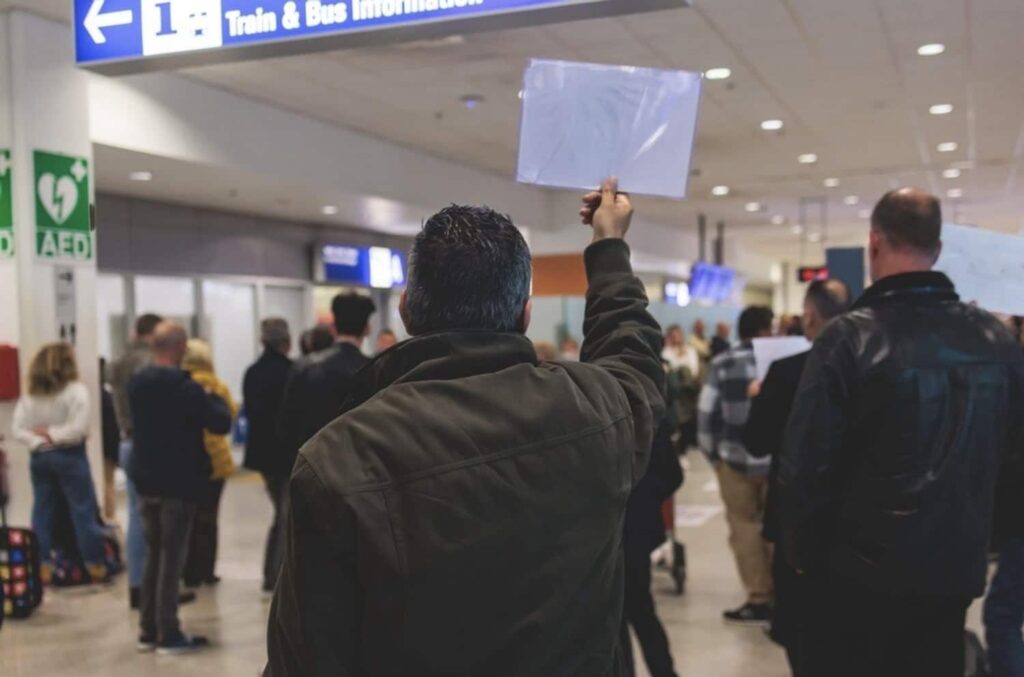 How to Get from CDG Airport to Paris: A man in a dark jacket holding up a clear plastic folder, likely containing a sign or nameplate, in a busy airport arrival hall. Passengers are walking by, with directional signs for train and bus information visible overhead.