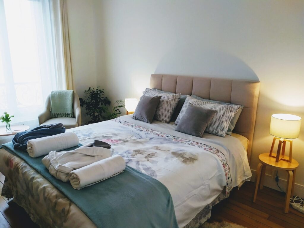airbnbs in paris: Inviting bedroom with natural light from a large window, featuring a plush double bed with a soft blue blanket and floral print, flanked by a cozy armchair with a throw and a wooden bedside table with a warm lamp. Fresh towels are neatly placed on the bed, creating a welcoming atmosphere for guests.