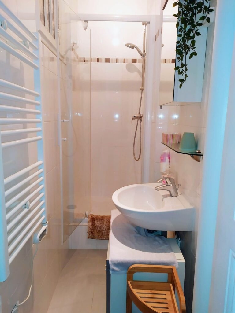 airbnbs in paris: Compact and clean bathroom interior with glossy white tiles, featuring a glass shower cubicle with a handheld shower head, a white basin on a pedestal with a towel draped over it, and a wooden bathroom stool. A heated towel rail and greenery add touches of comfort and freshness to the space.