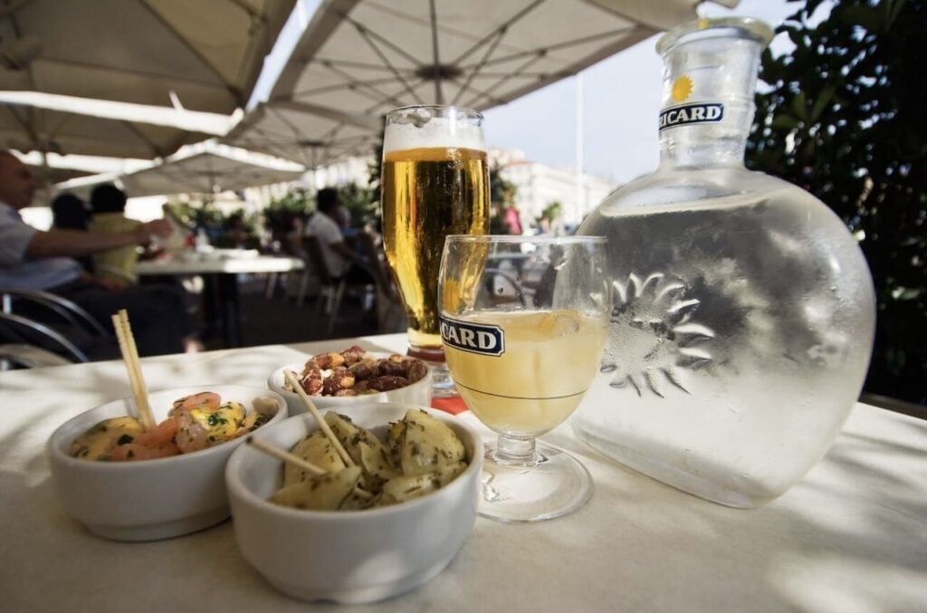 Al fresco dining scene with a bottle of Ricard pastis and a glass filled with the beverage beside a tall beer glass, accompanied by bowls of seasoned olives and nuts on a table under a patio umbrella.