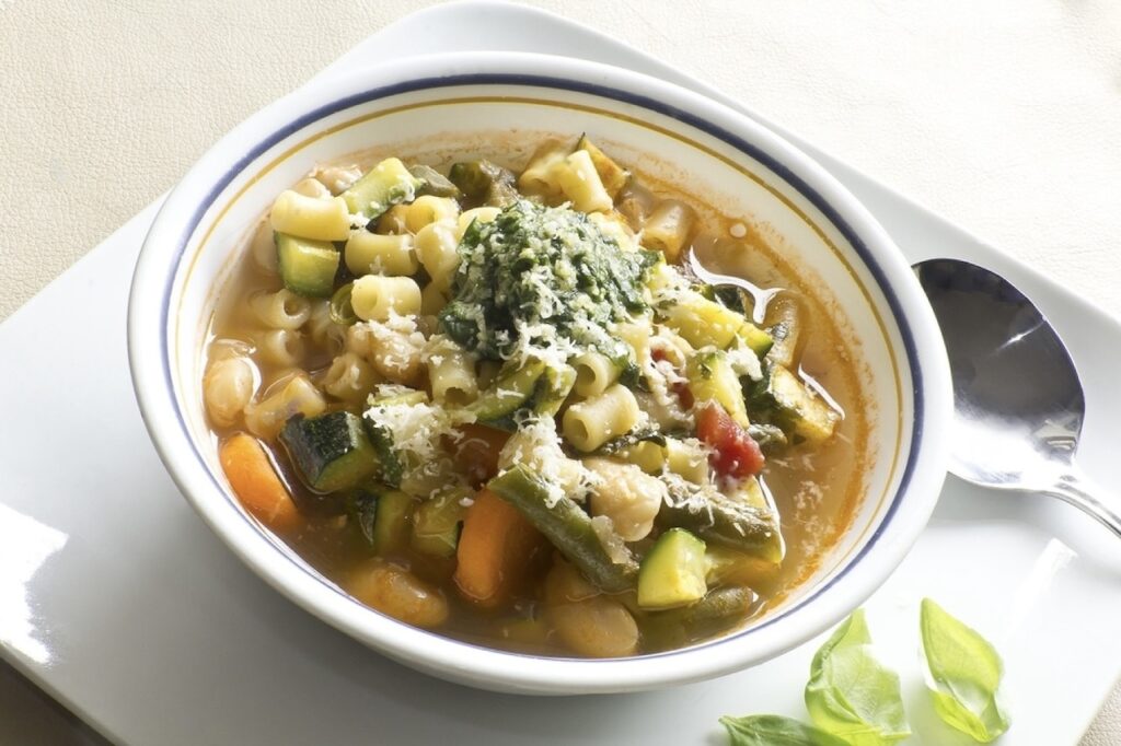 Marseille Food Guide: A hearty bowl of minestrone soup with mixed vegetables, beans, pasta, and a dollop of pesto on top, served with grated cheese, and accompanied by a spoon on a white plate, suggesting a wholesome and nourishing meal.