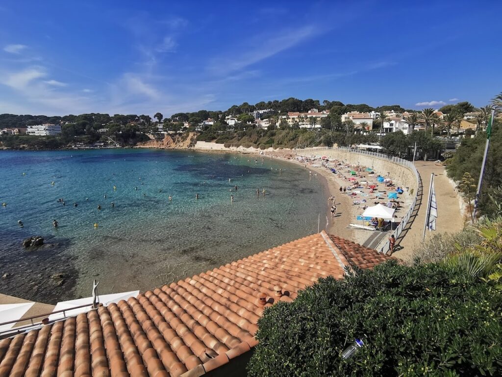 A scenic view of Portissol beach in Sanary-sur-Mer, with people enjoying the sun on the sandy shore and swimming in the clear blue waters, bordered by greenery and Mediterranean-style houses under a vivid blue sky.