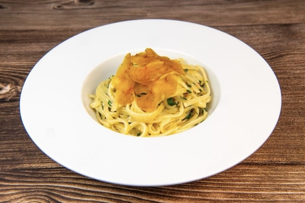 A plate of creamy fettuccine pasta garnished with herbs and topped with crispy potato chips, served on a white plate against a dark wood background.