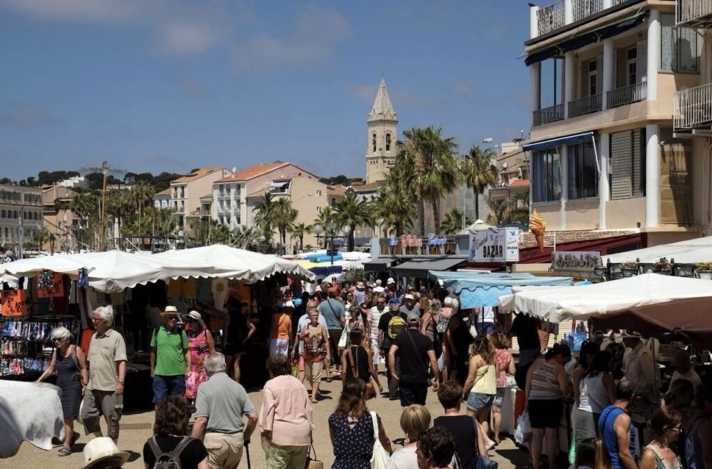 A bustling Provençal market in Sanary-sur-Mer, crowded with visitors browsing through stalls under white canopies, surrounded by palm trees and local architecture, with a historic church tower rising in the background under a clear blue sky.