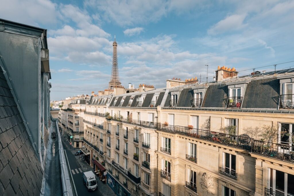 airbnbs in paris: Breathtaking view from a Parisian top-floor apartment, showcasing the iconic Eiffel Tower in the distance, with classic Haussmann-style buildings lining the street below under a clear blue sky.