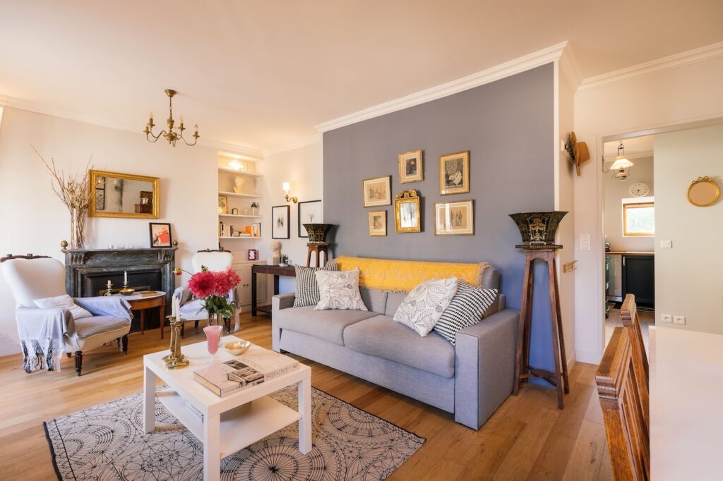 airbnbs in paris: Elegant and spacious living room with a cozy gray sofa adorned with patterned pillows, an antique fireplace and mirror, decorative art on a blue accent wall, and natural light streaming in, creating a welcoming Parisian home atmosphere.