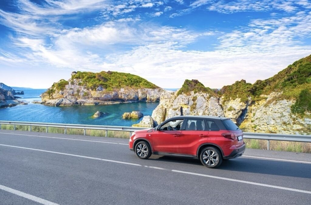 Renting a Car in France: A red SUV driving on a coastal road with a stunning view of rocky cliffs and a serene blue sea under a wispy clouded sky, evoking a sense of adventure and the beauty of seaside travel.