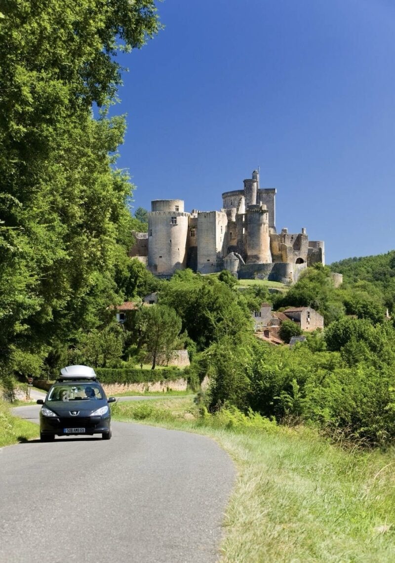 A scenic road leading to a majestic medieval castle surrounded by lush greenery under a clear blue sky. A car with a roof box is driving towards the castle, highlighting a mix of historical and modern travel.