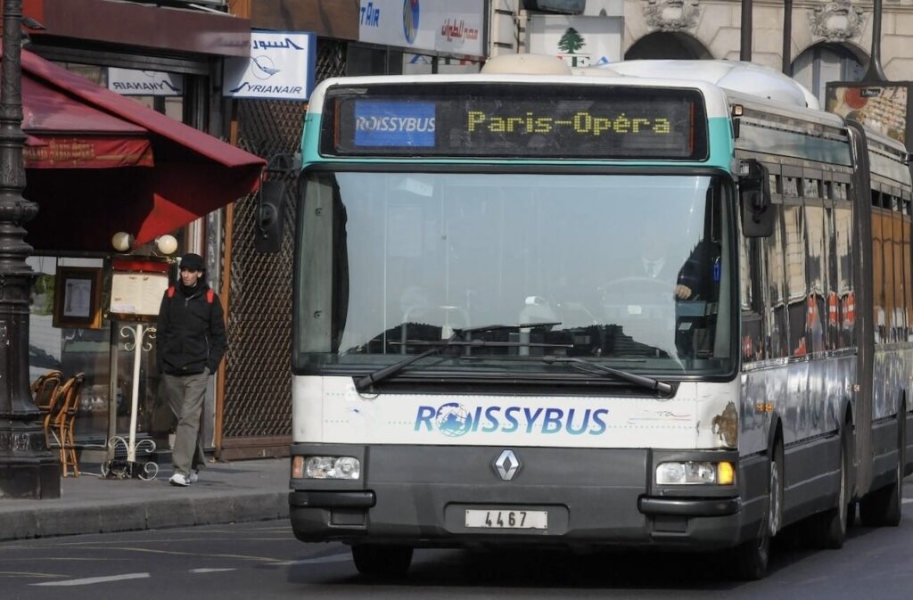 How to Get from CDG Airport to Paris: A Roissybus with 'Paris-Opera' displayed on its destination board is parked on a busy street, with various commercial signs and a pedestrian in casual attire standing nearby.
