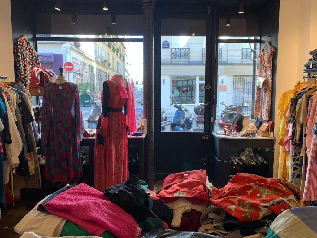 Vintage Shops in Paris: Inside view from Rose Market Vintage, with a glance through the open door onto a Parisian street. The shop interior is adorned with a vibrant selection of vintage dresses and assorted clothing items, reflecting the charm and fashion of bygone eras.