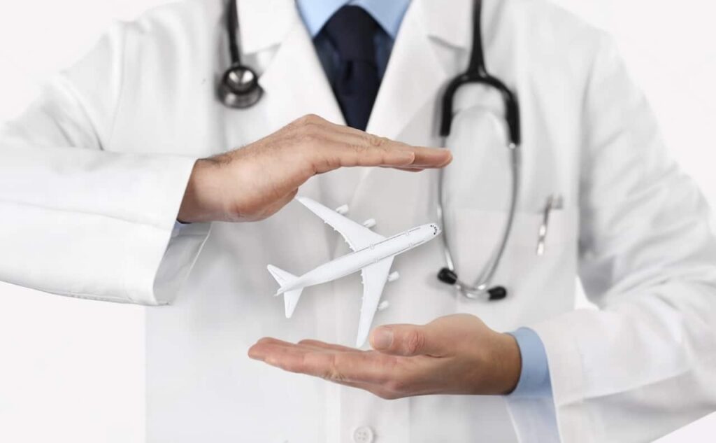 A doctor in a white coat and stethoscope is holding a miniature airplane between his hands, suggesting a concept of medical travel or healthcare services related to air travel and transport.