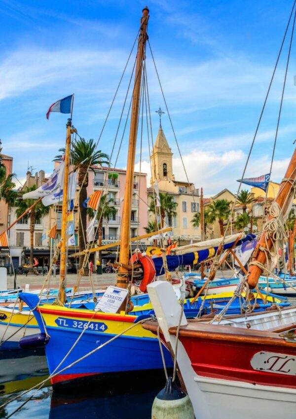 Colorful traditional fishing boats docked in the harbor of Sanary-sur-Mer, France, with the town's historic buildings and church steeple in the background against a clear blue sky.