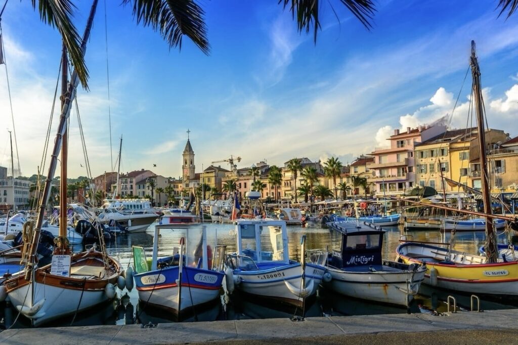 Traditional wooden boats moored in the harbor of Sanary-sur-Mer, Provence, with the town's picturesque buildings and church tower in the background, framed by palm leaves under a serene blue sky with soft clouds.