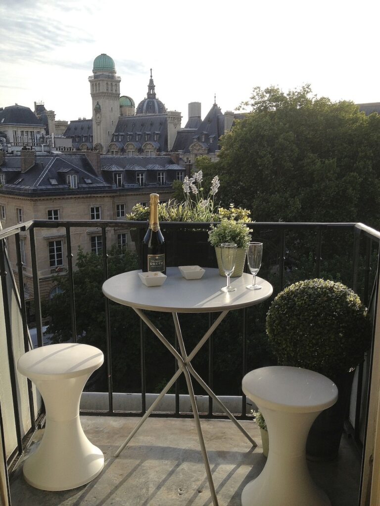 airbnbs in paris: Quaint balcony set up with a romantic view of Parisian rooftops and historic architecture, featuring a small round table with a bottle of champagne, two flutes, and a blooming plant, alongside modern white stools. The scene captures the essence of a relaxing Paris evening.