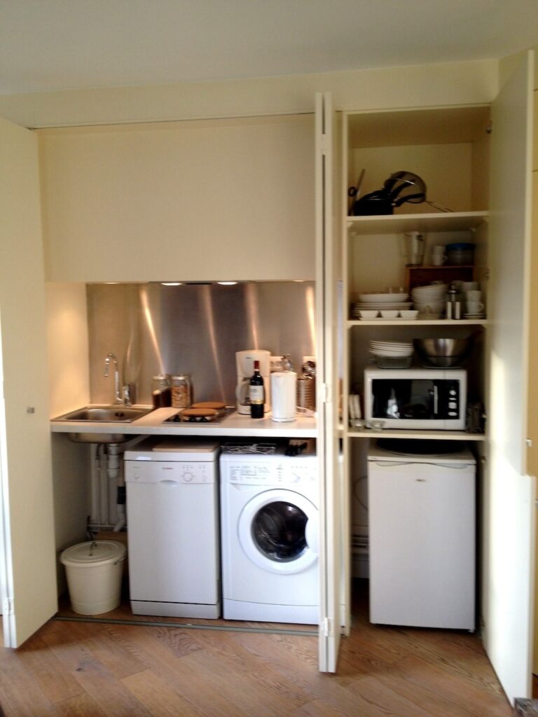 airbnbs in paris: Compact kitchenette neatly tucked into a closet space with open doors, revealing a stainless steel sink, white dishwasher, washing machine, and a small refrigerator. Shelves above hold a kettle, microwave, and an array of dishes and cups, making efficient use of the limited space.