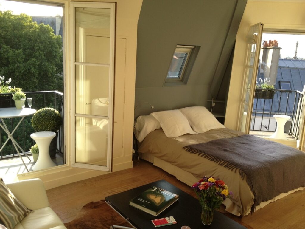 airbnbs in paris: Bright and sunny studio apartment with an open French door leading to a balcony with a view of lush greenery. The room features a cozy bed with a taupe coverlet and white pillows, a modern white sofa, and a coffee table with a bouquet of colorful flowers, creating a welcoming and airy atmosphere.