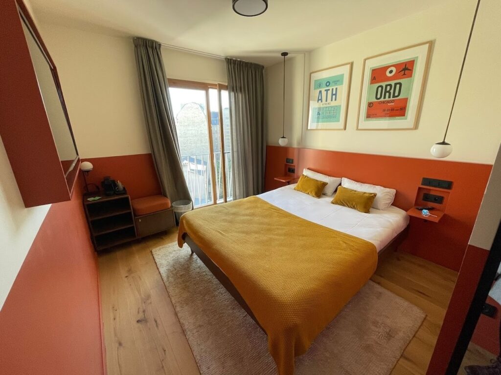 gay hotels in Paris: Bright and colorful hotel room with a bold terracotta accent wall, complemented by retro travel-themed artwork. The cozy space features a comfortable bed with a golden yellow throw, matching pillows, and a balcony letting in natural light, creating an inviting and warm atmosphere for guests.