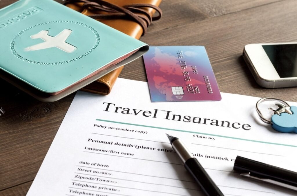 A close-up view of a travel insurance form ready to be filled out, with a pen on top, alongside a passport holder, credit card, smartphone, and keychain, all laid out on a wooden surface as part of travel preparations.