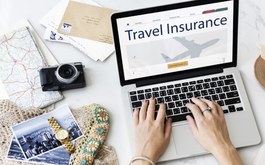 Travel Insurance for France: Hands typing on a laptop displaying a 'Travel Insurance' webpage, with related travel items like a map, camera, postcards, and a wristwatch spread around, depicting the planning phase of a journey with a focus on the importance of securing travel insurance.