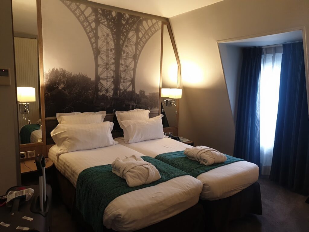 A Paris-themed hotel room with twin beds covered in white linens and emerald green throws, accented by towels shaped like swans. A large mural of the Eiffel Tower spans the wall behind the beds, while natural light filters in through the blue curtains, enhancing the room's intimate and romantic atmosphere.