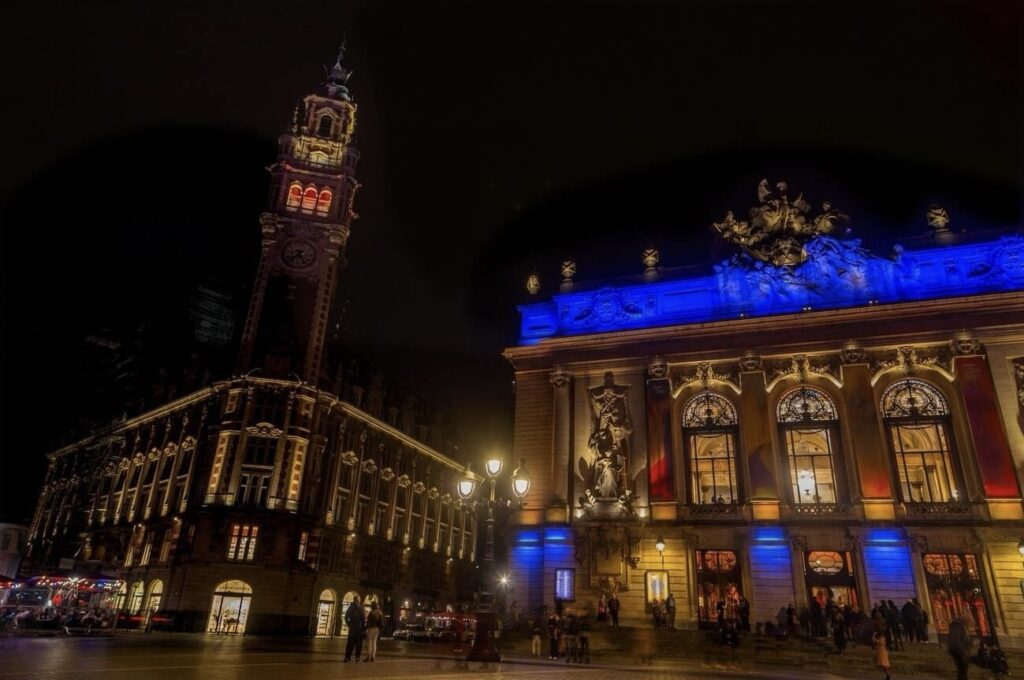 Night view of the illuminated Lille Opera House with its grand clock tower standing tall next to the vibrant, blue-lit facade, as people gather and stroll in the foreground.