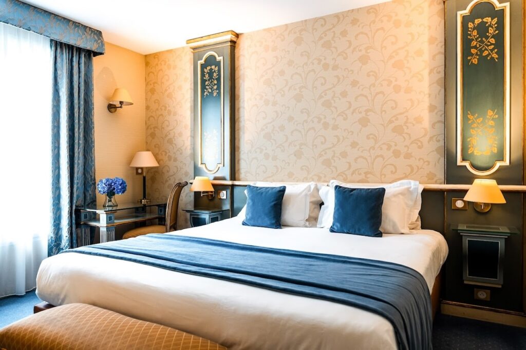gay hotels in Paris: An elegantly appointed hotel room with a regal cream and gold wallpaper, luxurious blue velvet pillows on a large bed, and ornate framed mirrors adding a touch of classic French decor. A vibrant bouquet of blue flowers on a vintage desk enhances the room's sophisticated charm.