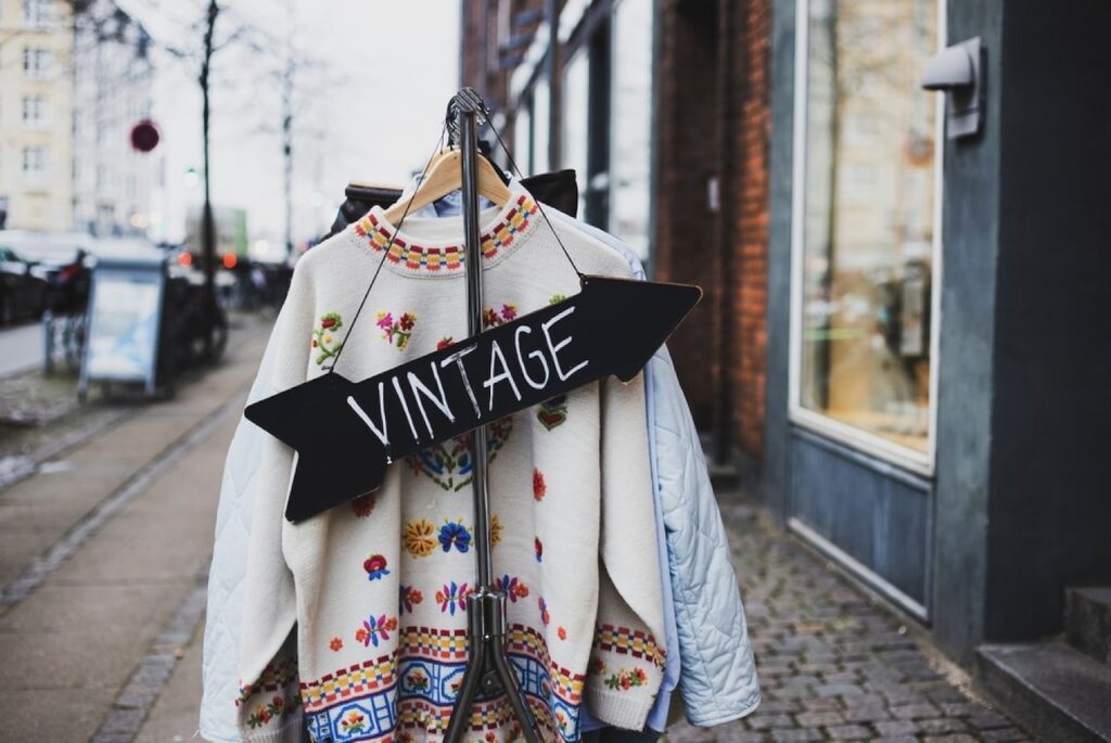 Vintage Shops in Paris: A cozy vintage sweater with floral embroidery hangs on a rack outdoors, with a black arrow sign pointing left towards the word 'VINTAGE', inviting shoppers into the store on a city street with a European feel.