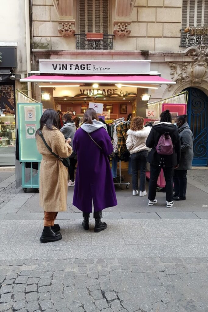 Vintage Shops in Paris: Shoppers in warm coats browse through a street-side vintage clothing rack outside 'VINTAGE By Ramin' store, with the bustling activity of a Parisian sidewalk scene encapsulating the allure of city life and second-hand fashion finds.
