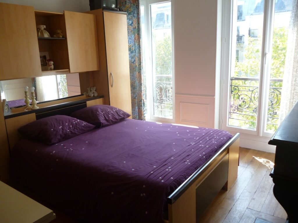 airbnbs in paris: A cozy bedroom featuring a large bed with a purple bedspread, natural light pouring in through French windows with intricate railings, flanked by built-in wooden cabinets and a reflective mirror, creating a warm and inviting space.
