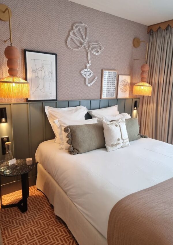 Elegant hotel room with white bedding, green pillows, and herringbone-patterned wallpaper. Unique line art and sculptural pieces add artistic flair, while ambient lighting creates a warm, inviting atmosphere.