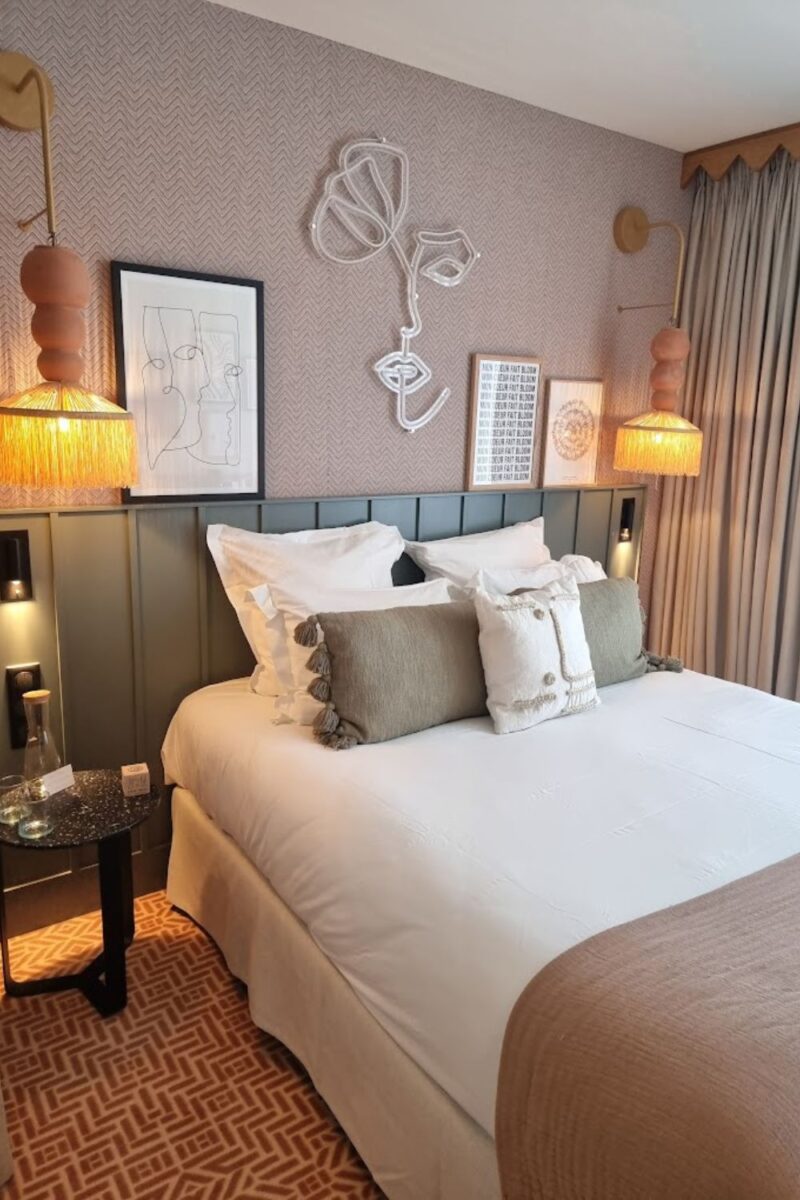 Elegant hotel room with white bedding, green pillows, and herringbone-patterned wallpaper. Unique line art and sculptural pieces add artistic flair, while ambient lighting creates a warm, inviting atmosphere.