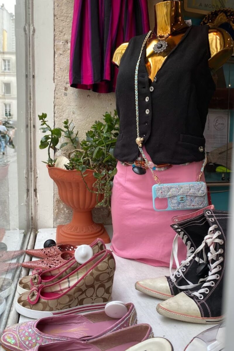 A whimsical window display features a gold mannequin torso dressed in a black vest and pink skirt, accompanied by an eclectic mix of vintage shoes and a bedazzled purse, set against the backdrop of a charming Paris street.