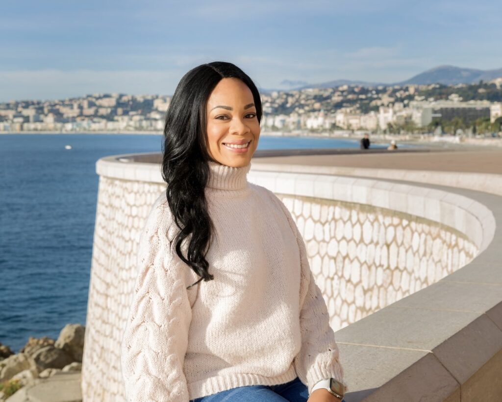 A smiling woman in a cozy sweater sits by the seafront promenade, with the panoramic view of Nice's coastline behind her, capturing the essence of a relaxed 'Nice in one day' experience.