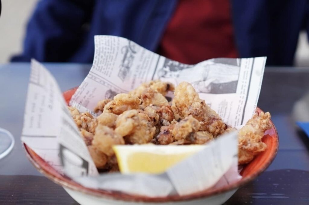Marseille Food Guide: A bowl of crispy fried baby squid, also known as 'chipirones fritos', served in a red bowl lined with newspaper, garnished with a lemon wedge, with a blurred background of a person at a dining table, emphasizing a typical tapas experience.