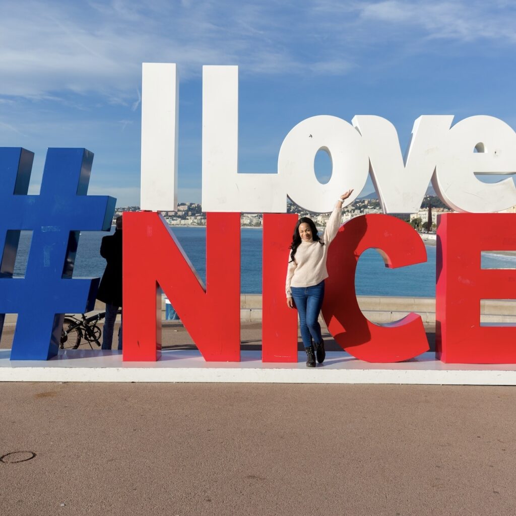 A smiling woman poses in front of the iconic #ILoveNice sign on the Promenade des Anglais, a popular photo spot for visitors completing the 'Nice in one day' challenge.