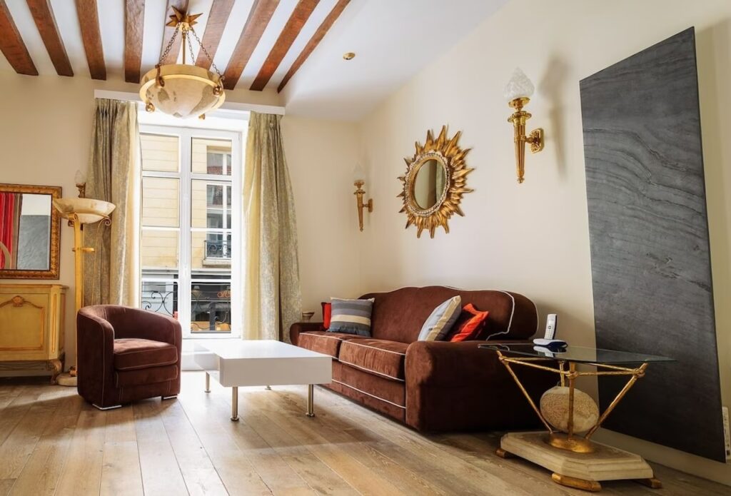 airbnbs in paris: Luxuriously appointed living room with a plush brown velvet sofa and matching armchair, elegant gold-framed sunburst mirror, classic sconce lighting, and exposed wooden ceiling beams, alluding to the historical charm of a Parisian apartment.
