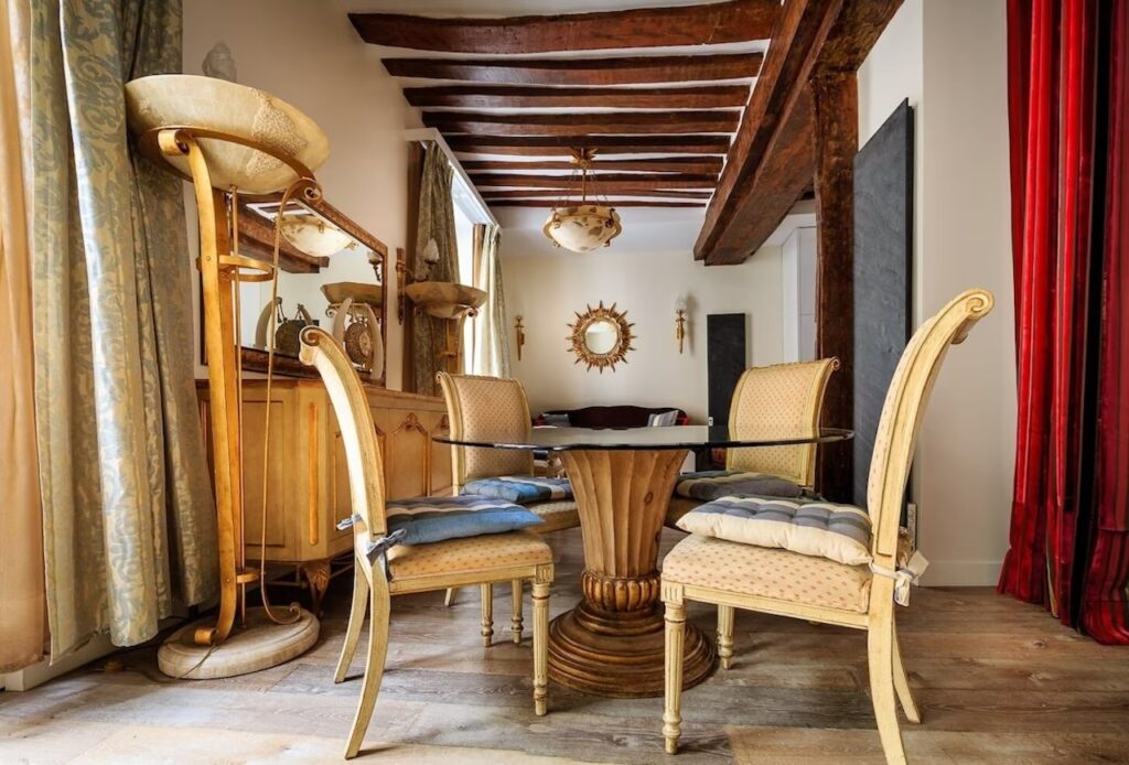 airbnbs in paris: A charming dining area with antique flair, featuring a circular wooden table, elegant upholstered chairs, a vintage mirror, and a sunburst wall accent, all under the rustic ambiance of exposed ceiling beams and warm drapery.
